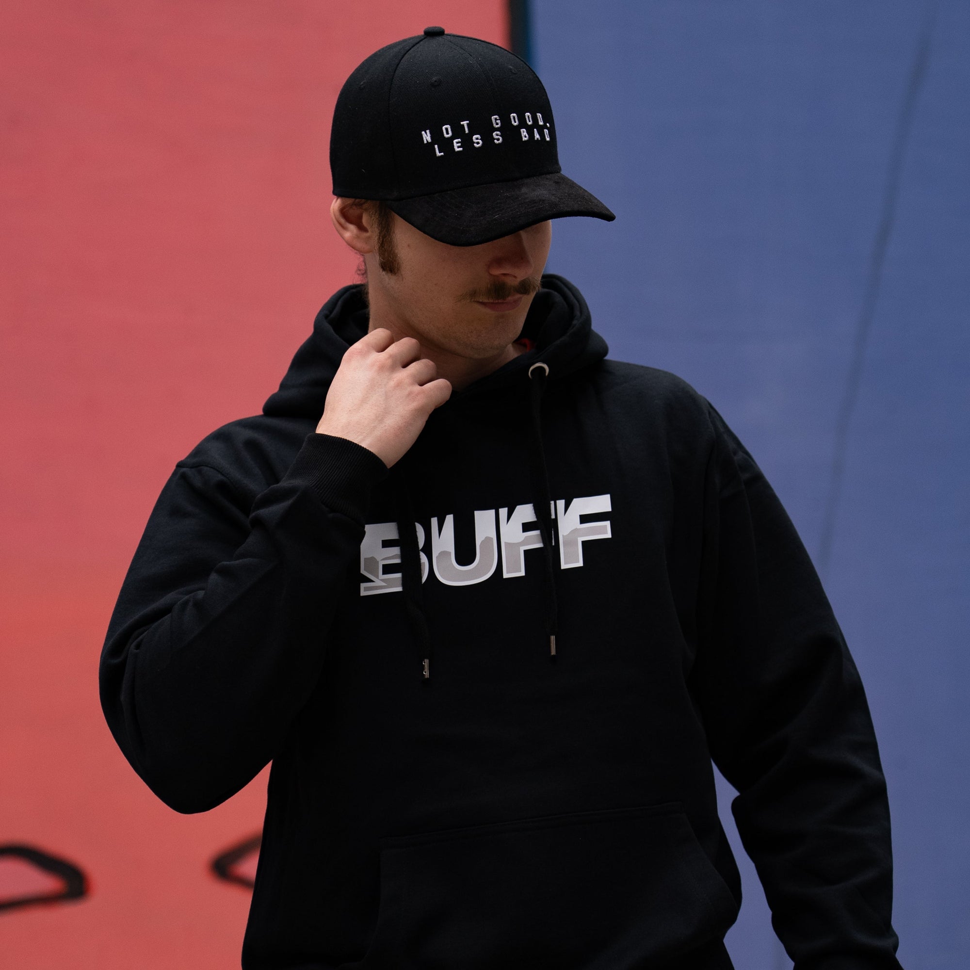 Person wearing a black hoodie with 'BUFF' branding and a black cap against a red and blue background.