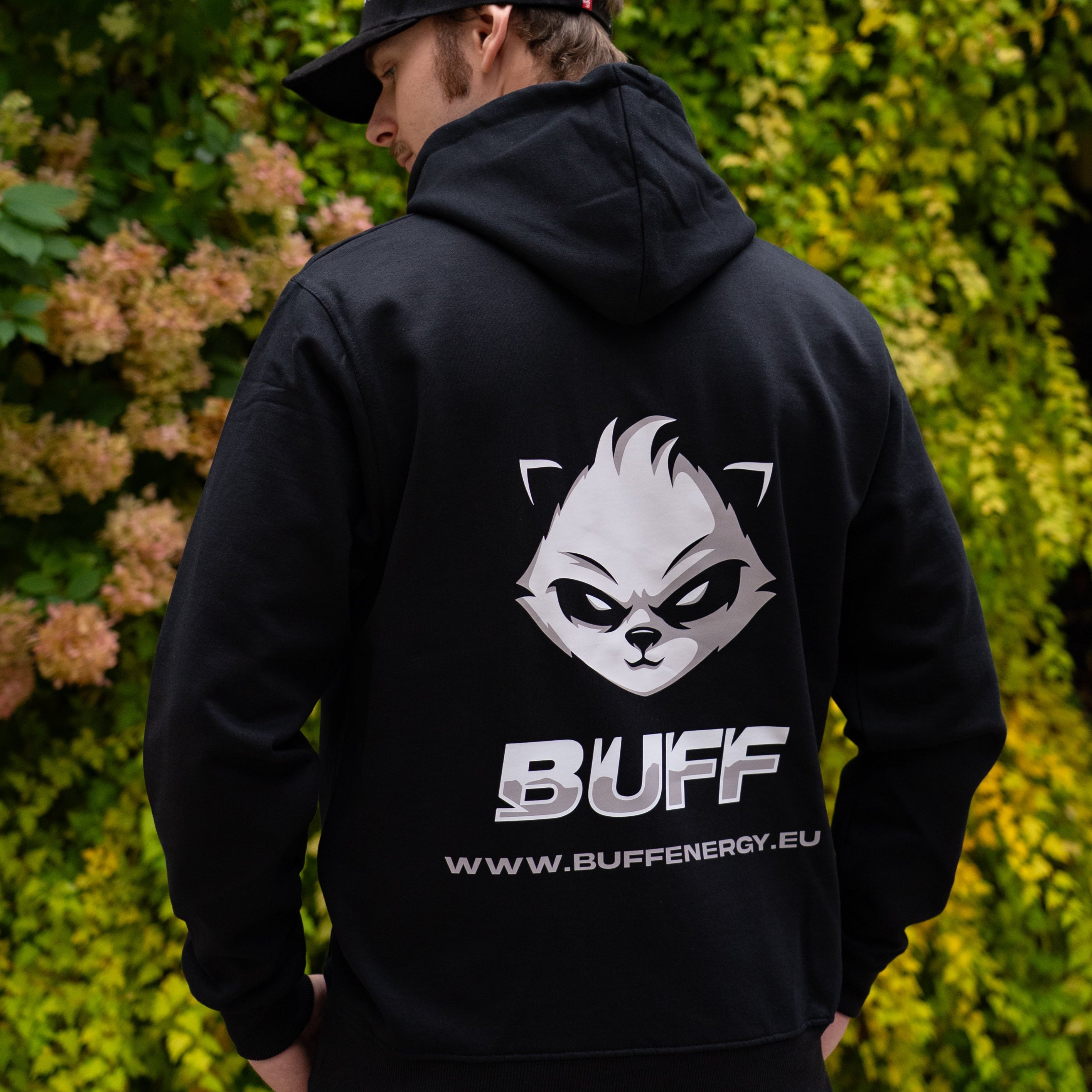 Person wearing a black hoodie with a logo and text on the back, standing in front of green foliage.
